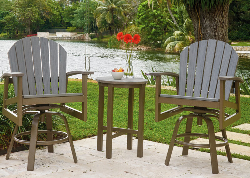 Two rustic polymer Adirondack chairs face each other by a small table with a bowl of fruit and a vase of red flowers, set against a lush, garden backdrop with a calm river.