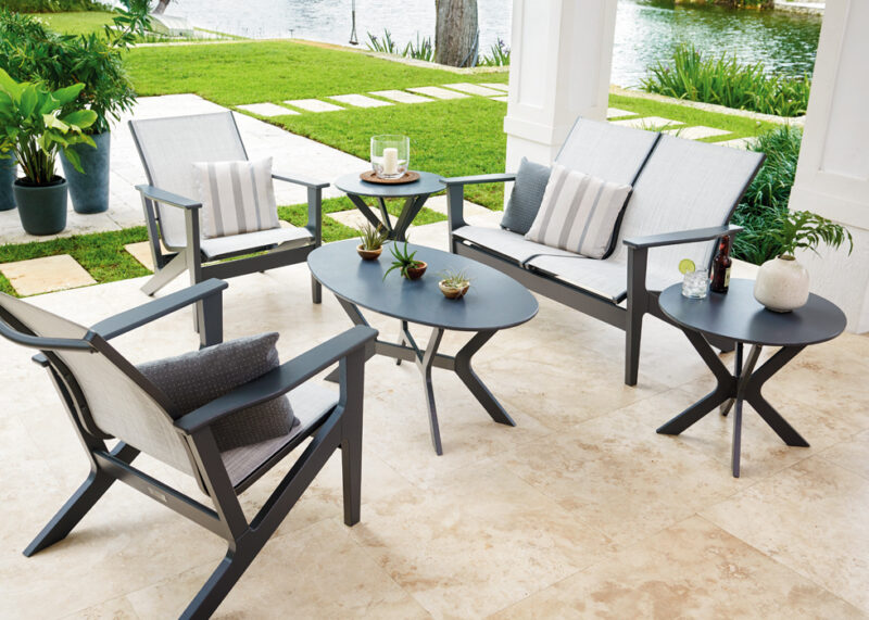 Outdoor patio furniture set including a sofa, two armchairs, and two round tables on a tiled patio overlooking a lawn and a water body. decorated with cushions and a small plant on the table.