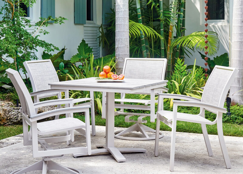 An outdoor patio setup with a rustic polymer top table and white chairs, featuring a plate of oranges and a glass of juice, surrounded by lush green tropical plants.