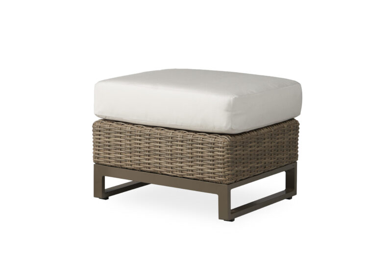 A beige, wicker ottoman with a thick, white cushion on top, set against a plain white background. the ottoman features a sturdy metal frame visible at the base.