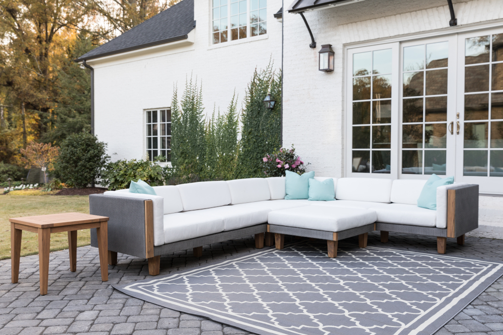 An outdoor seating area with a modular Catalina white sofa arranged in an L-shape, accessorized with light blue pillows, on a gray patterned rug. A wooden side table and lush greenery