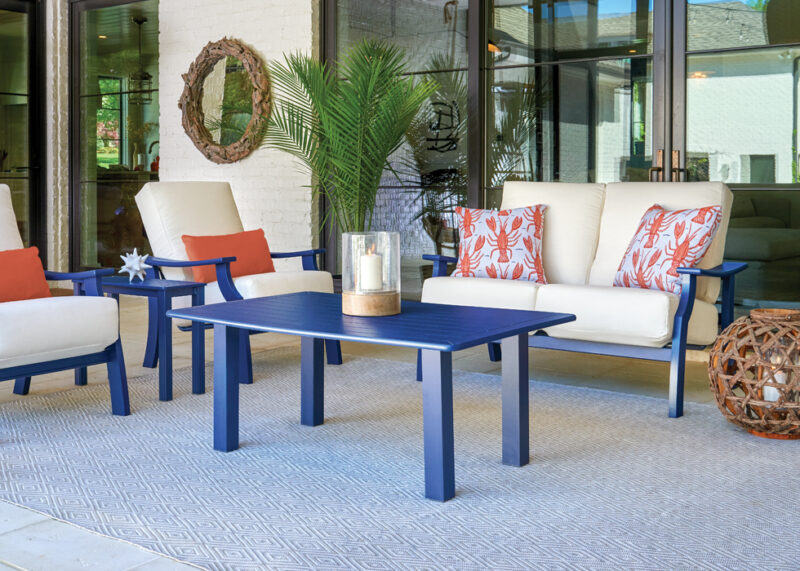A stylish outdoor seating area with two white sofas adorned with orange and white patterned cushions, flanked by two red armchairs, a blue center table, all on a textured rug under a covered patio.