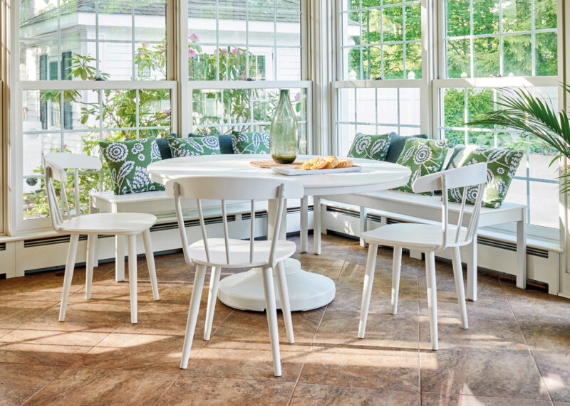 Bright sunroom dining area with a white round table and chairs, patterned cushions, and large windows showing green trees outside. a vase and fruit on the table add a homely touch.