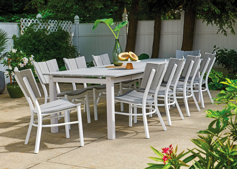 An outdoor dining area featuring a long rustic polymer top table with several chairs set in a lush garden, accompanied by a decorative plant and a plate of fruit on the table.