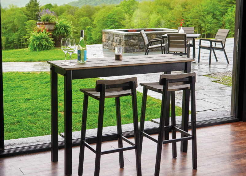 Outdoor patio seating area with a rustic polymer top table and two stools, wine glasses, and a bottle on the table, overlooking a lush green landscape with a stone wall and trees.