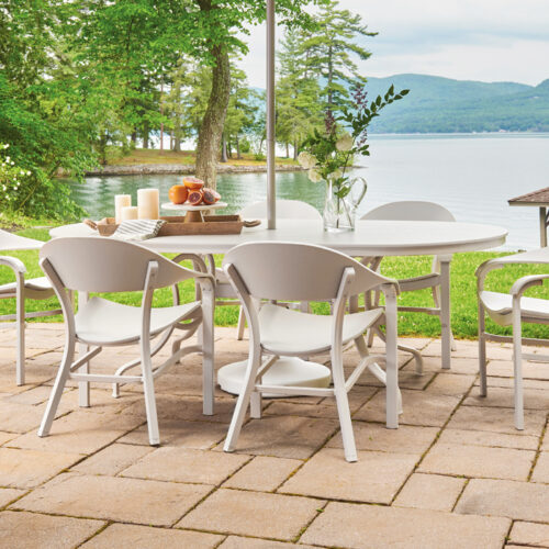 Outdoor patio by a lake with two white dining tables and several chairs, set for a meal, surrounded by trees with distant mountains visible under a cloudy sky.