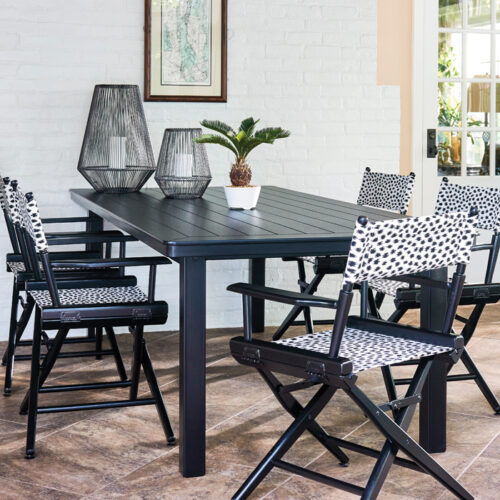 Modern dining area with a black table and chairs featuring white polka dot cushions. the room includes white brick walls, large windows, and decorative plants.