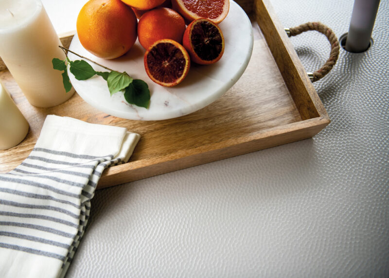 A wooden tray holds a white plate with fresh oranges and blood oranges, next to a striped cloth and a bottle, on a textured light surface.