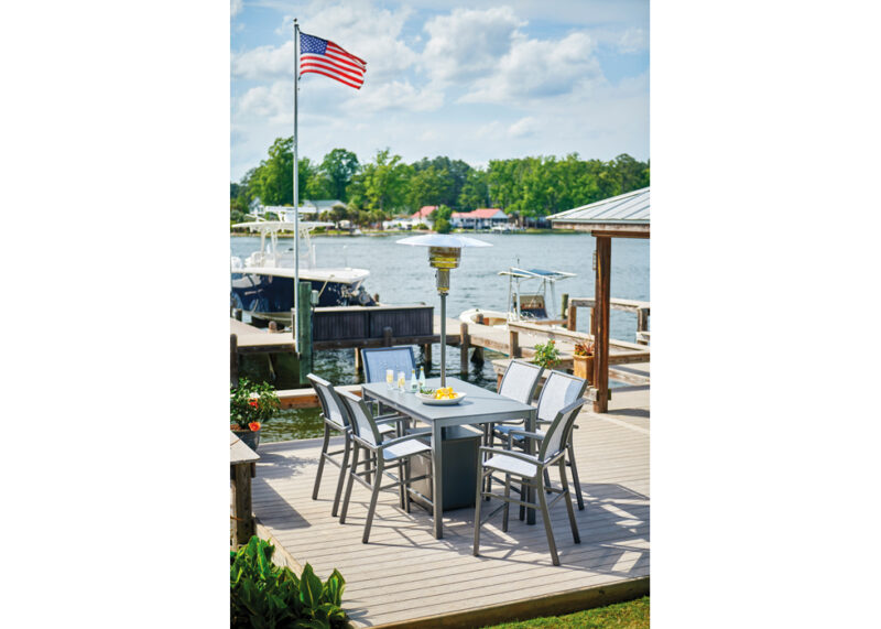 An outdoor dining setup on a waterfront dock featuring a table set for breakfast with an american flag waving above, overlooking boats and a lake under a clear sky.