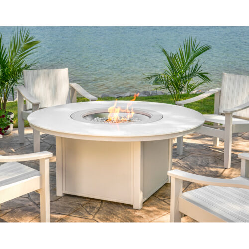 A serene outdoor seating area by the lake featuring an Origins Top Fire Table surrounded by comfortable white chairs, with lush green plants and a clear view of the water.