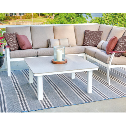 Outdoor patio overlooking a lake with a modern beige sectional sofa with red pillows, a white coffee table, striped gray rug, and potted plants, creating a serene setting.