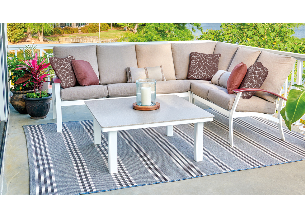 Outdoor patio overlooking a lake with a modern beige sectional sofa with red pillows, a white coffee table, striped gray rug, and potted plants, creating a serene setting.