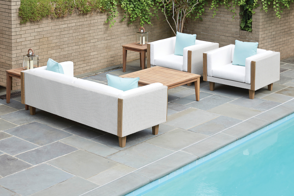 Outdoor patio with modern white woven Catalina furniture, including a sofa and two chairs with light blue cushions, beside a swimming pool. Wooden tables with lamps are placed next to the seating.