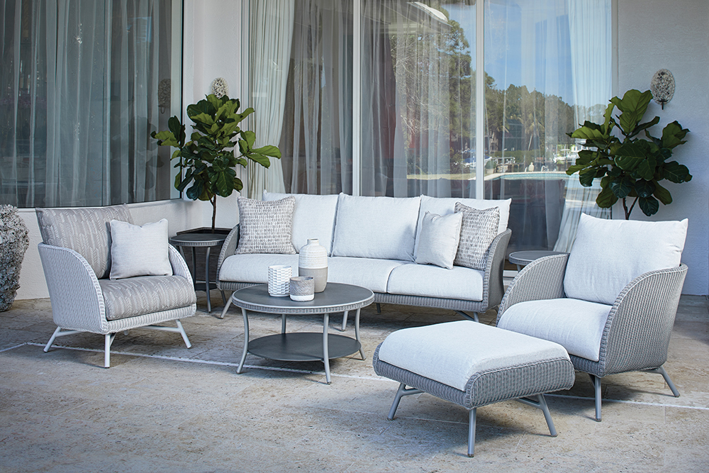 Outdoor patio exuding the essence of modernity with wicker furniture featuring white cushions, including a sofa, chairs, and a chaise lounge, arranged around a round coffee table near sleek glass doors.
