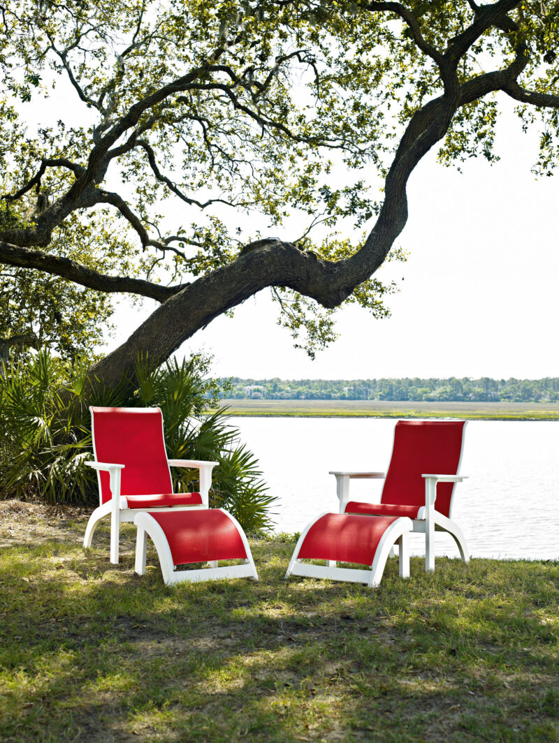 Two red patio chairs facing a tranquil lake, shaded by an overarching tree on a sunny day, suggesting a peaceful outdoor retreat.