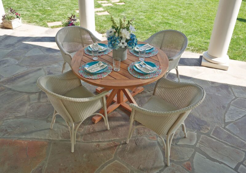 Outdoor dining setup with a round wooden table and four wicker chairs on a stone patio. the table is set with plates, cutlery, and a vase with blue flowers.