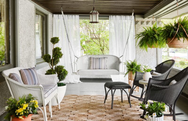 A cozy, well-decorated porch with a hanging bench, chairs, lush plants, curtains, and a textured rug. the space exudes a relaxing atmosphere with a blend of modern and rustic elements.