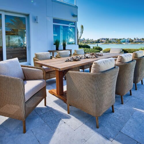 Luxurious outdoor dining area with a wooden table and cushioned wicker chairs on a tile floor, overlooking a serene waterfront view under a clear blue sky.