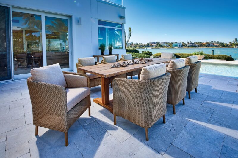 Luxurious outdoor dining area with a wooden table and cushioned wicker chairs on a tile floor, overlooking a serene waterfront view under a clear blue sky.
