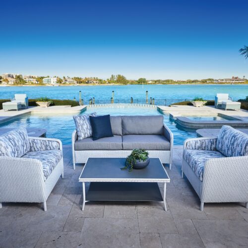 Luxurious outdoor patio by a tranquil blue lake with comfort seating including a sofa and armchairs, arranged around a white coffee table. clear skies and calm waters enhance the serene setting.