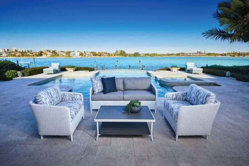 Luxurious outdoor patio by a tranquil blue lake with comfort seating including a sofa and armchairs, arranged around a white coffee table. clear skies and calm waters enhance the serene setting.