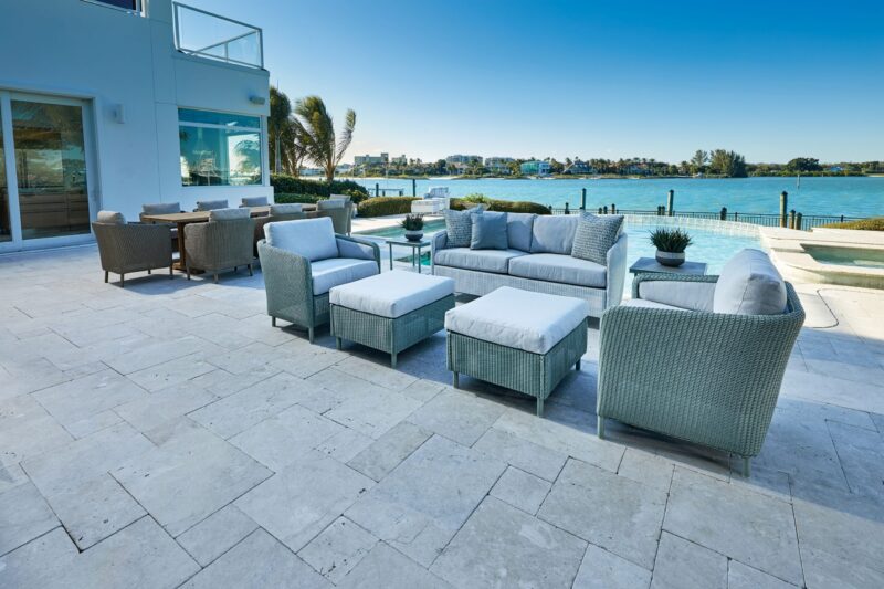 Outdoor patio furniture set consisting of grey wicker sofas with light blue cushions and a glass-top coffee table, situated on a stone terrace overlooking a tranquil blue bay.