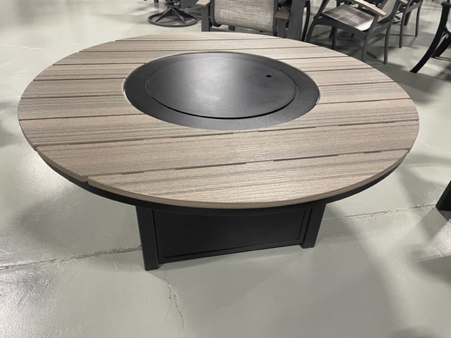 Round wooden table with a central black metal fire pit, surrounded by Newport Adirondack chairs, displayed on a grey concrete floor in a showroom setting.