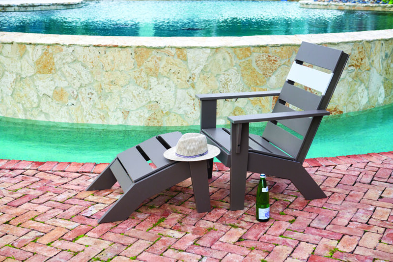 A gray adirondack chair on a brick patio beside an outdoor pool, equipped with a sun hat on the armrest and a bottle of beer next to it.