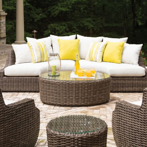 An outdoor seating area featuring a curved wicker sofa with white and yellow cushions, a round glass-topped coffee table with drinks, surrounded by matching chairs on a stone patio.