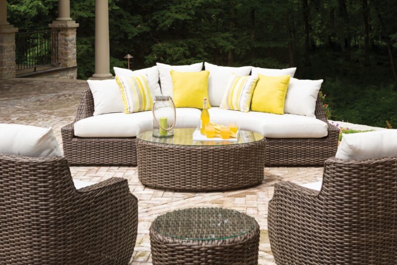 An outdoor seating area featuring a curved wicker sofa with white and yellow cushions, a round glass-topped coffee table with drinks, surrounded by matching chairs on a stone patio.