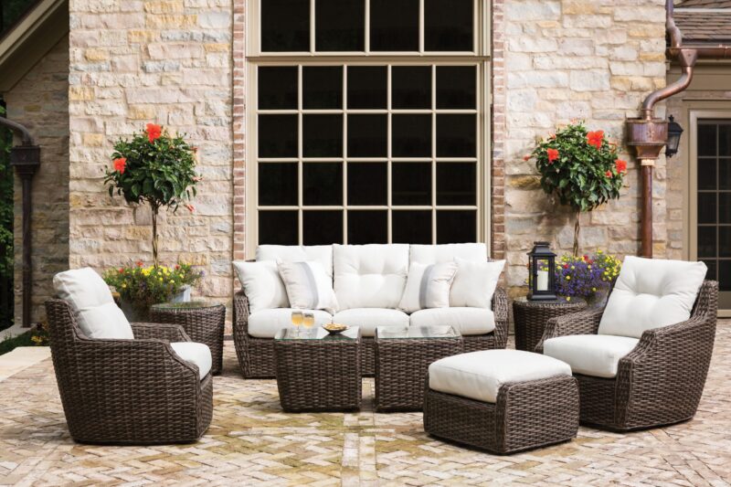 An outdoor seating area with a stone wall background featuring a large window. the set includes a sofa, chairs, and a table, all made of woven material with white cushions, and hanging plants.