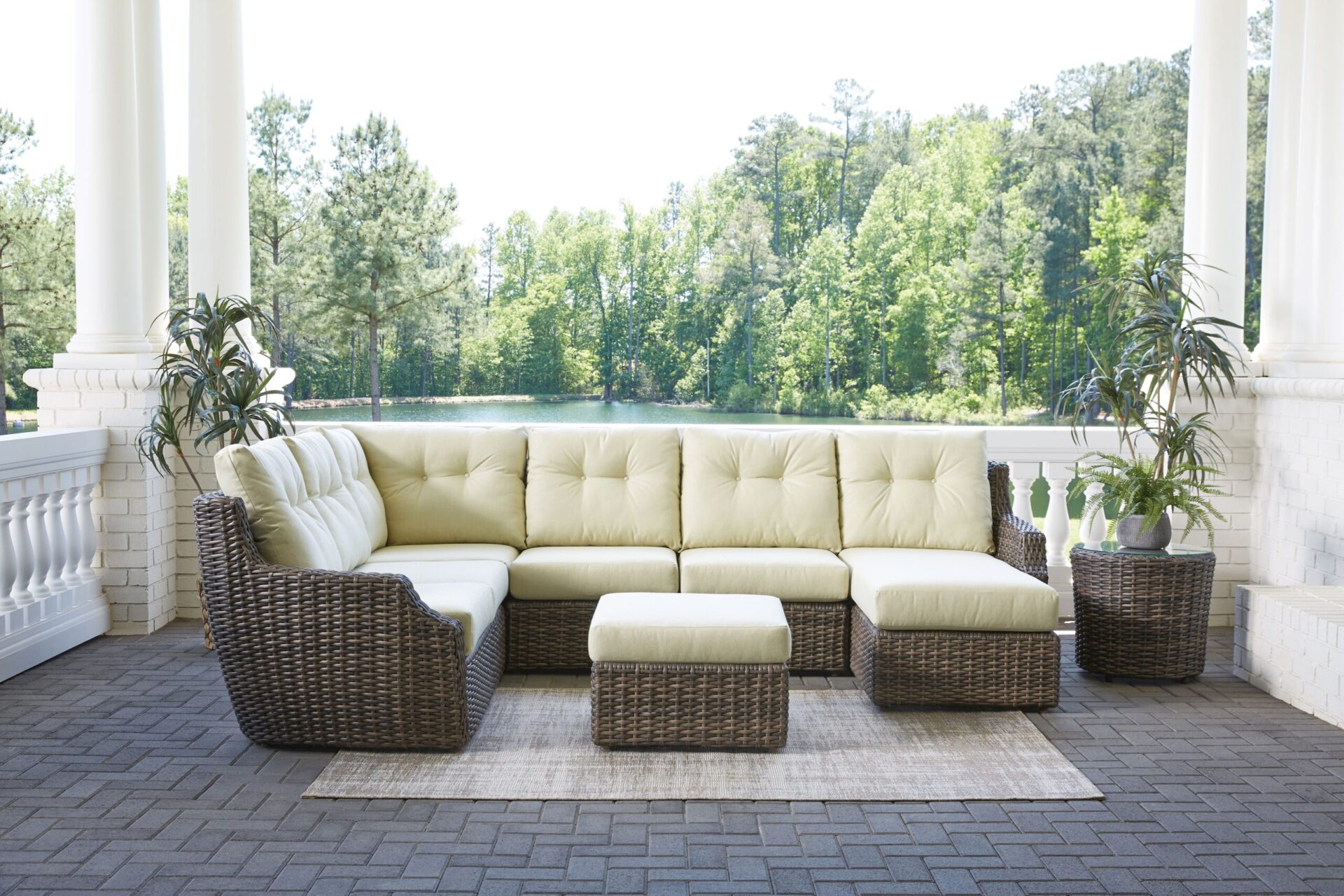 An elegant outdoor seating area on a tiled patio with a large beige modular sofa, two wicker chairs, and potted plants, surrounded by white columns overlooking lush greenery.