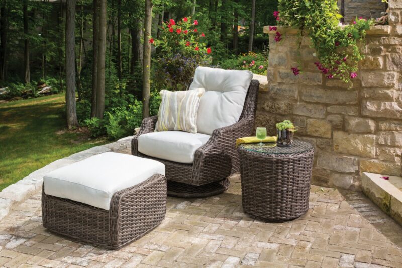 An outdoor patio with a rattan armchair and matching ottoman set on stone pavers, complemented by a round rattan side table. the setting is lush with greenery and blooming flowers.