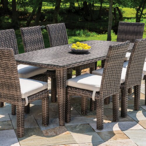 An outdoor dining set with six rattan chairs and a rectangular table, topped with a bowl of lemons, set on a stone patio surrounded by lush greenery.