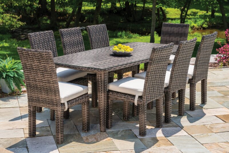 An outdoor dining set with six rattan chairs and a rectangular table, topped with a bowl of lemons, set on a stone patio surrounded by lush greenery.