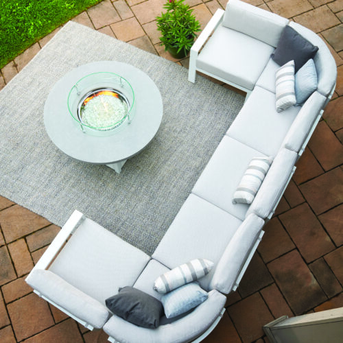 An aerial view of a stylish outdoor seating arrangement featuring a gray sectional sofa with white cushions, a round white coffee table on a rug, set on a brick patio surrounded by green grass.