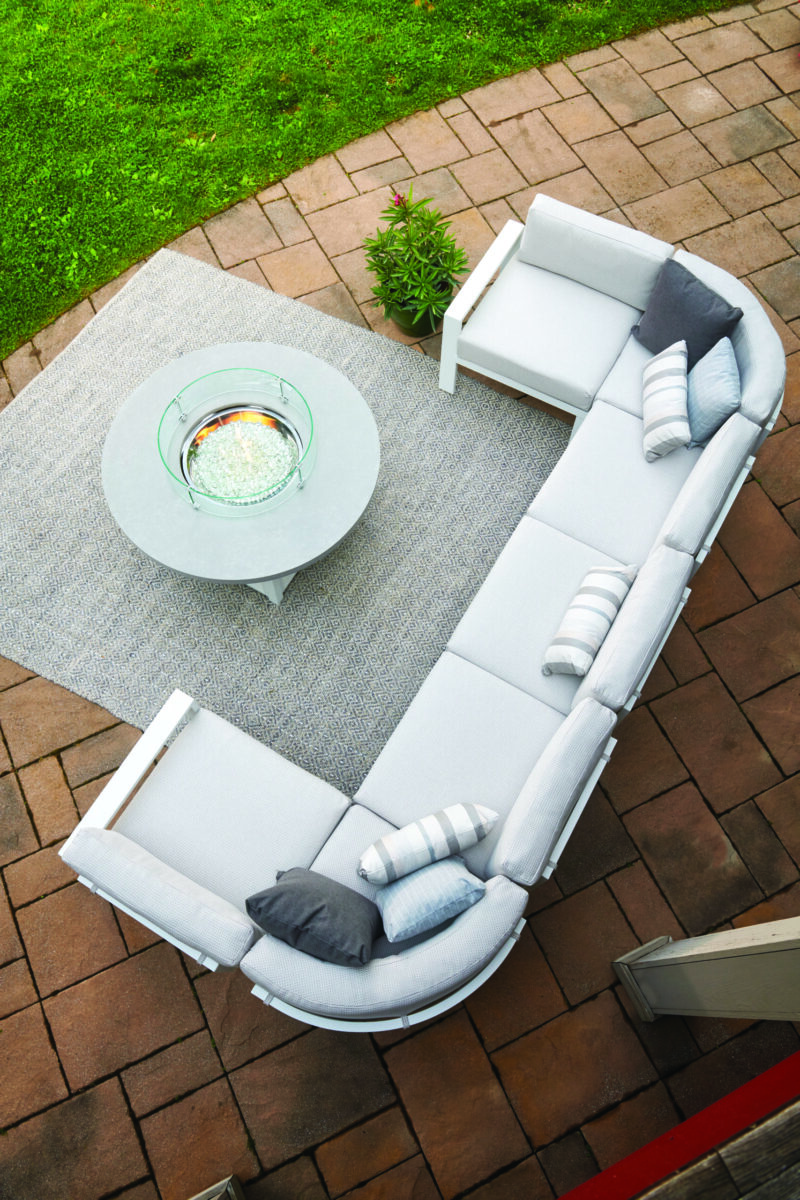 An aerial view of a stylish outdoor seating arrangement featuring a gray sectional sofa with white cushions, a round white coffee table on a rug, set on a brick patio surrounded by green grass.