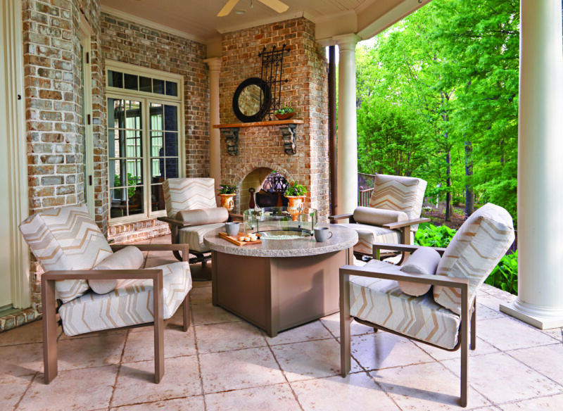 A cozy outdoor patio with a brick fireplace, elegant furniture including a round table and chairs, and a view of lush greenery.