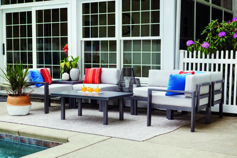 A stylish outdoor seating area by a pool with modern gray furniture, colorful cushions, and decorative plants, near a white picket fence and large windows.