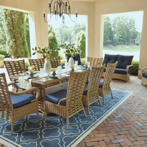 Outdoor dining setup under a covered patio with a chandelier, featuring a table set for six and two additional lounge chairs, surrounded by lush greenery.