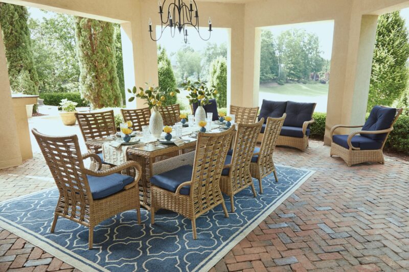 Outdoor dining setup under a covered patio with a chandelier, featuring a table set for six and two additional lounge chairs, surrounded by lush greenery.