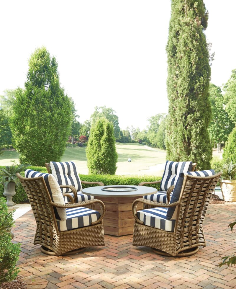 Four wicker chairs with blue and white striped cushions arranged around a circular table on a brick patio, overlooking a lush garden and golf course in the background.