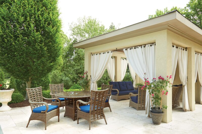 Elegant outdoor patio area with a dining set and a lounging space, featuring wicker chairs, blue cushions, flowing white curtains, and potted plants, all under a gazebo.