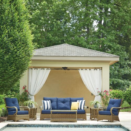 An elegant poolside cabana with a beige roof and draped white curtains, featuring a sectional sofa with blue cushions and striped pillows, flanked by potted plants.