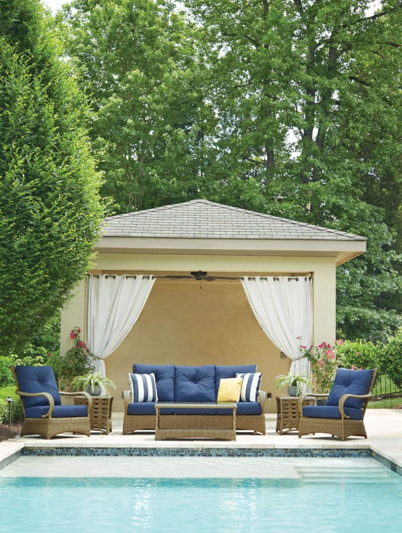 An elegant poolside cabana with a beige roof and draped white curtains, featuring a sectional sofa with blue cushions and striped pillows, flanked by potted plants.