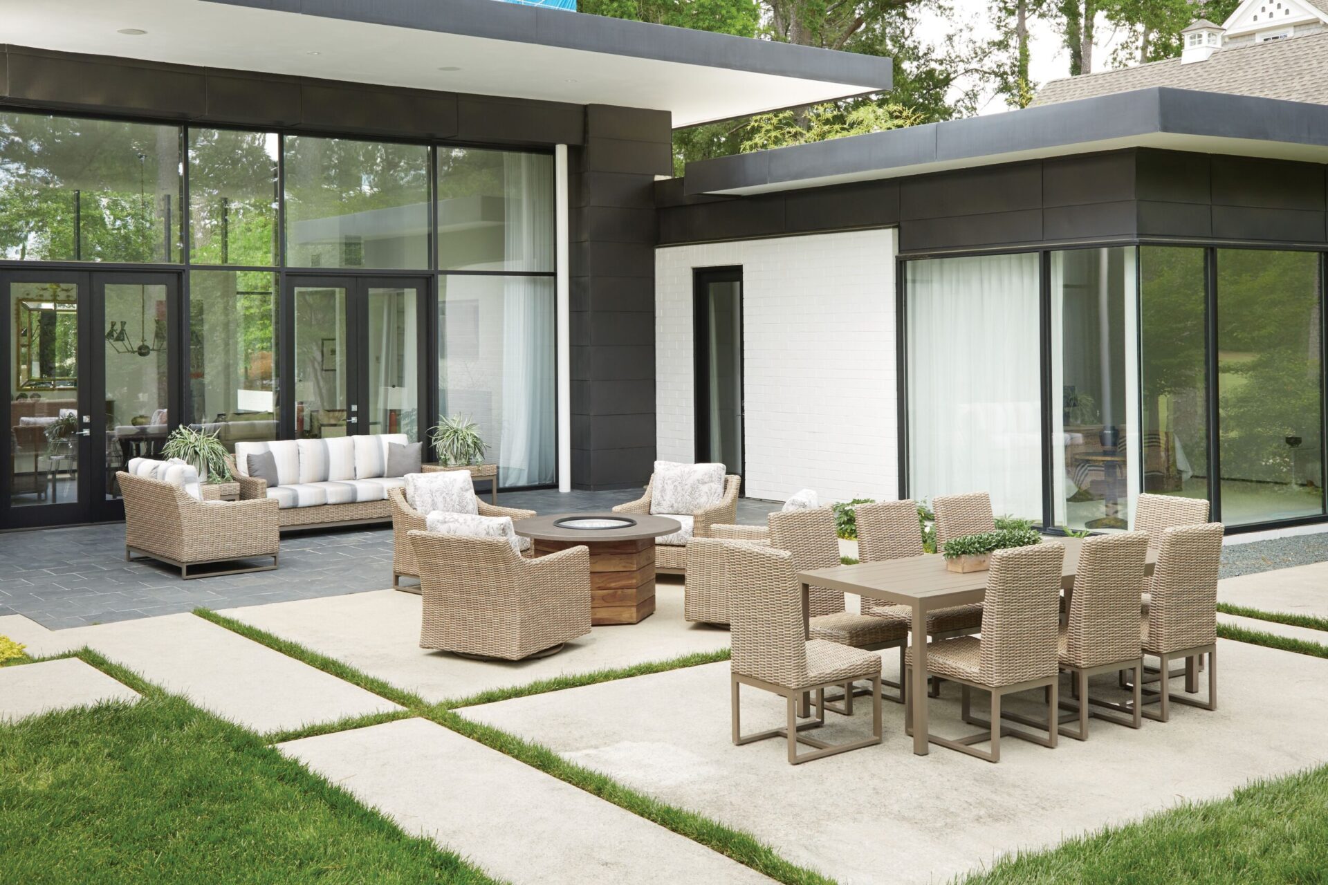 Modern patio area with Milan-inspired wicker furniture and a dining set on a manicured lawn, next to a contemporary house with large glass windows and white walls.