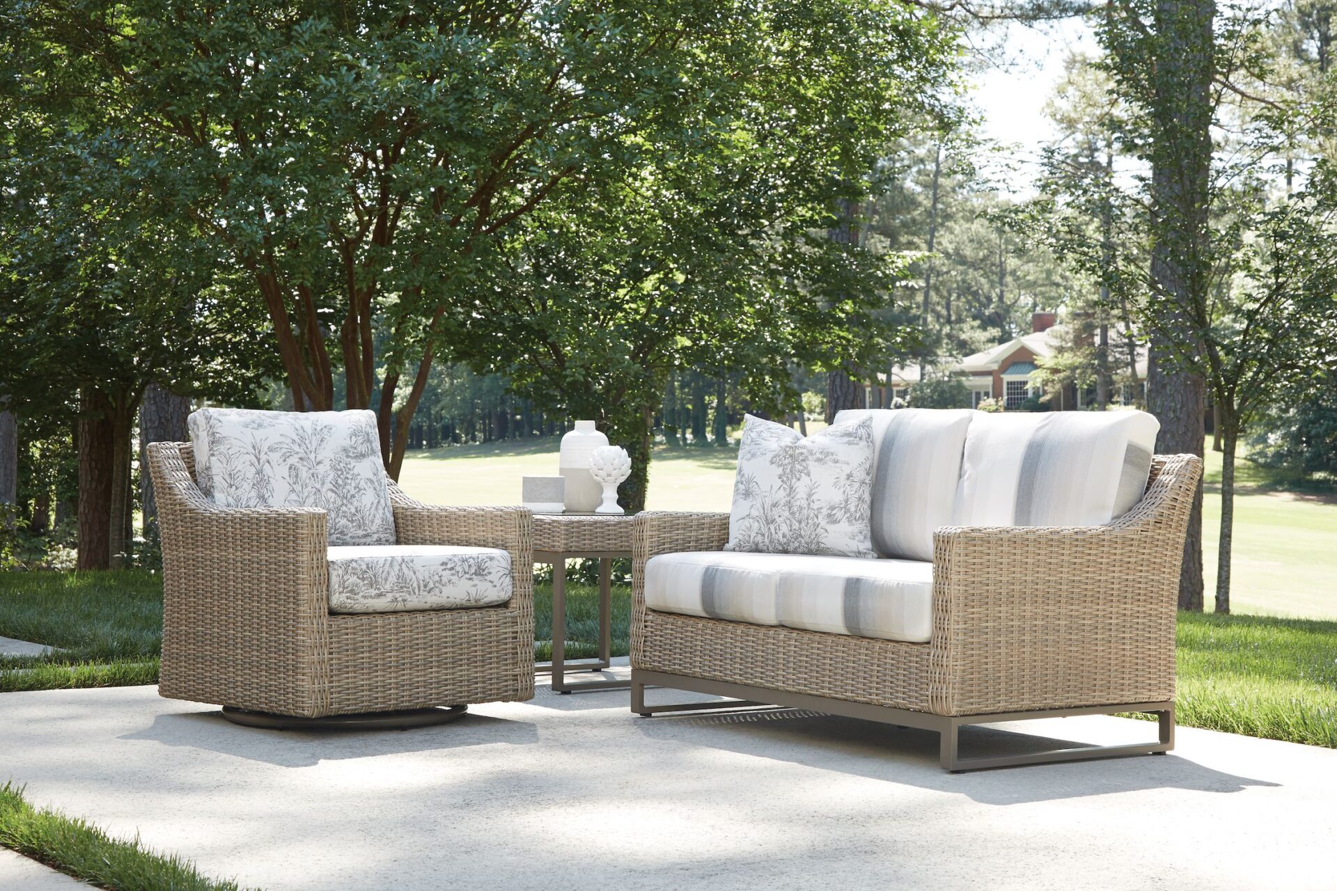 Two Milan-style wicker sofas with white cushions on a concrete patio surrounded by lush trees, creating a serene outdoor seating area.