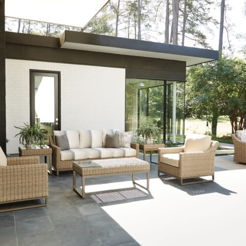 A modern patio inspired by Milanese design, with wicker furniture including two chairs, a sofa, and a coffee table, set against a white brick wall with a tall mirror, overlooking a sunny lawn