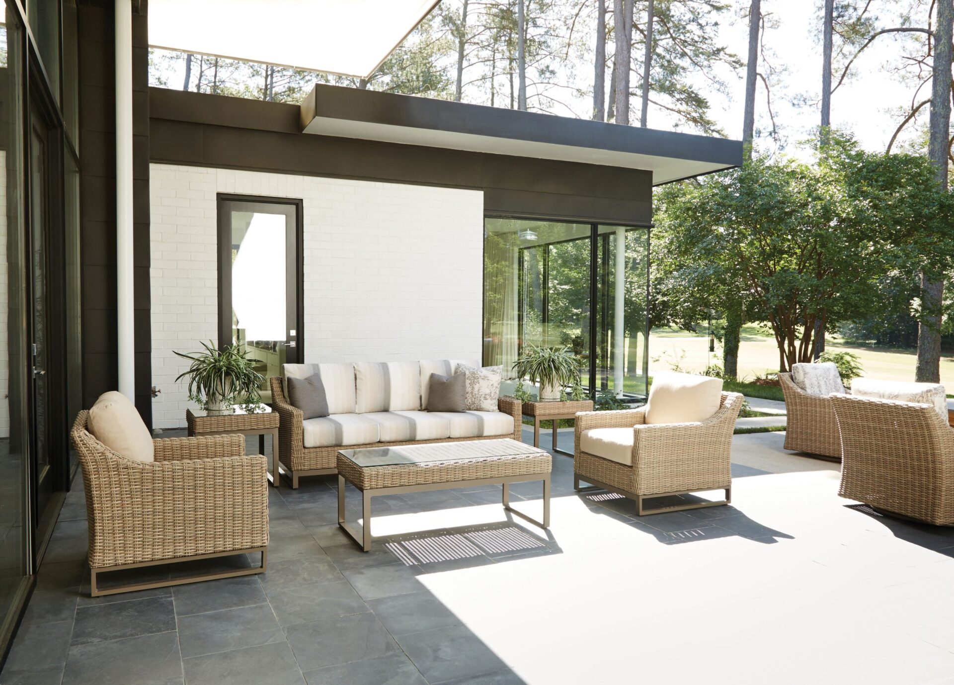 A modern patio inspired by Milanese design, with wicker furniture including two chairs, a sofa, and a coffee table, set against a white brick wall with a tall mirror, overlooking a sunny lawn