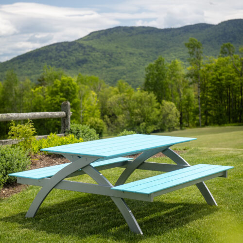A light blue picnic table on green grass with lush mountains in the background at Plymouth Bay under a partly cloudy sky.